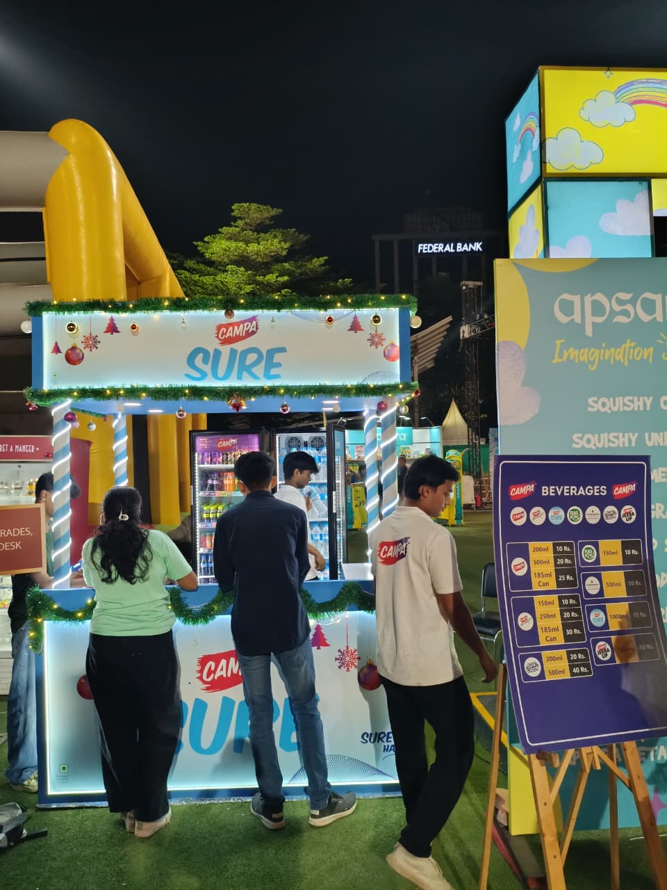 The Campa Sure hydration booth lit up at night, three guests served at the counter, a Christmas garland strung along the roofline and a yellow inflatable slide behind it