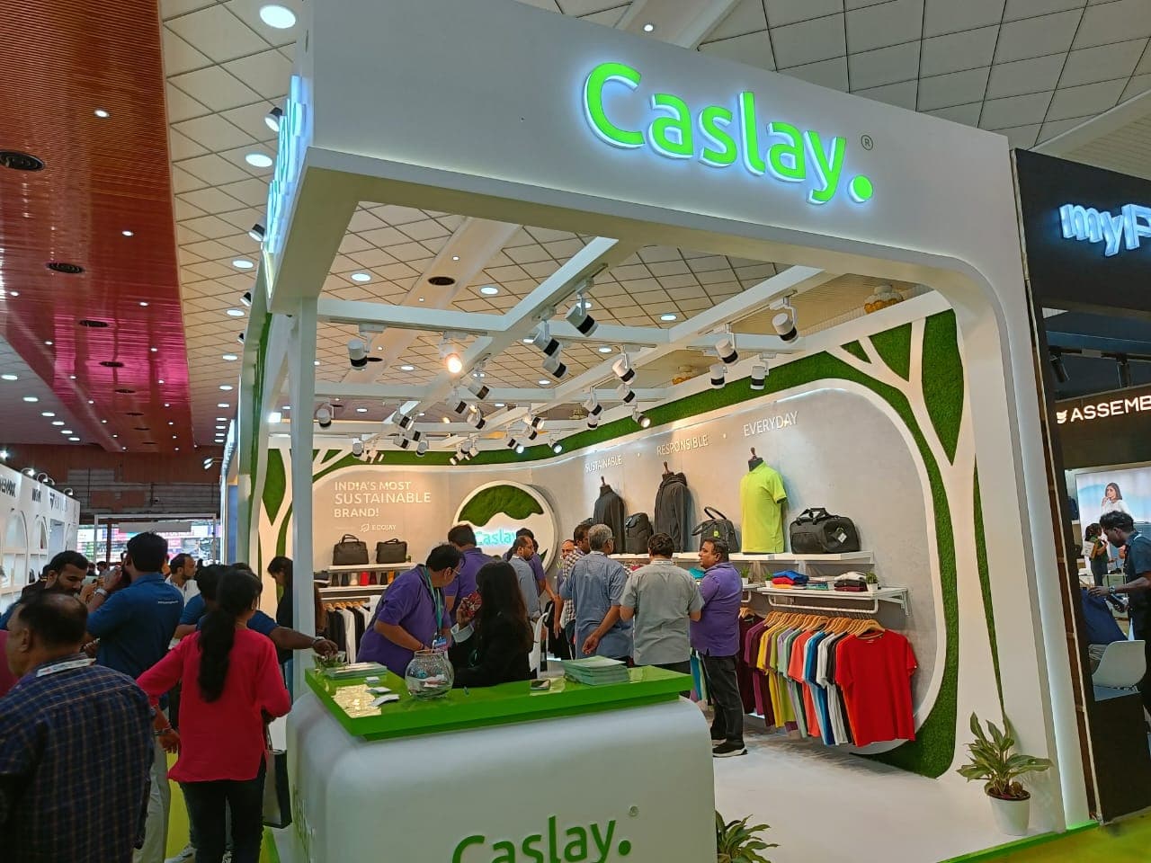 The Caslay exhibition booth crowded with visitors, staff in purple polo shirts assisting them at the counter, the 'India's Most Sustainable Brand!, powered by ECOJAY' badge and the tree-silhouette wall lit up behind them