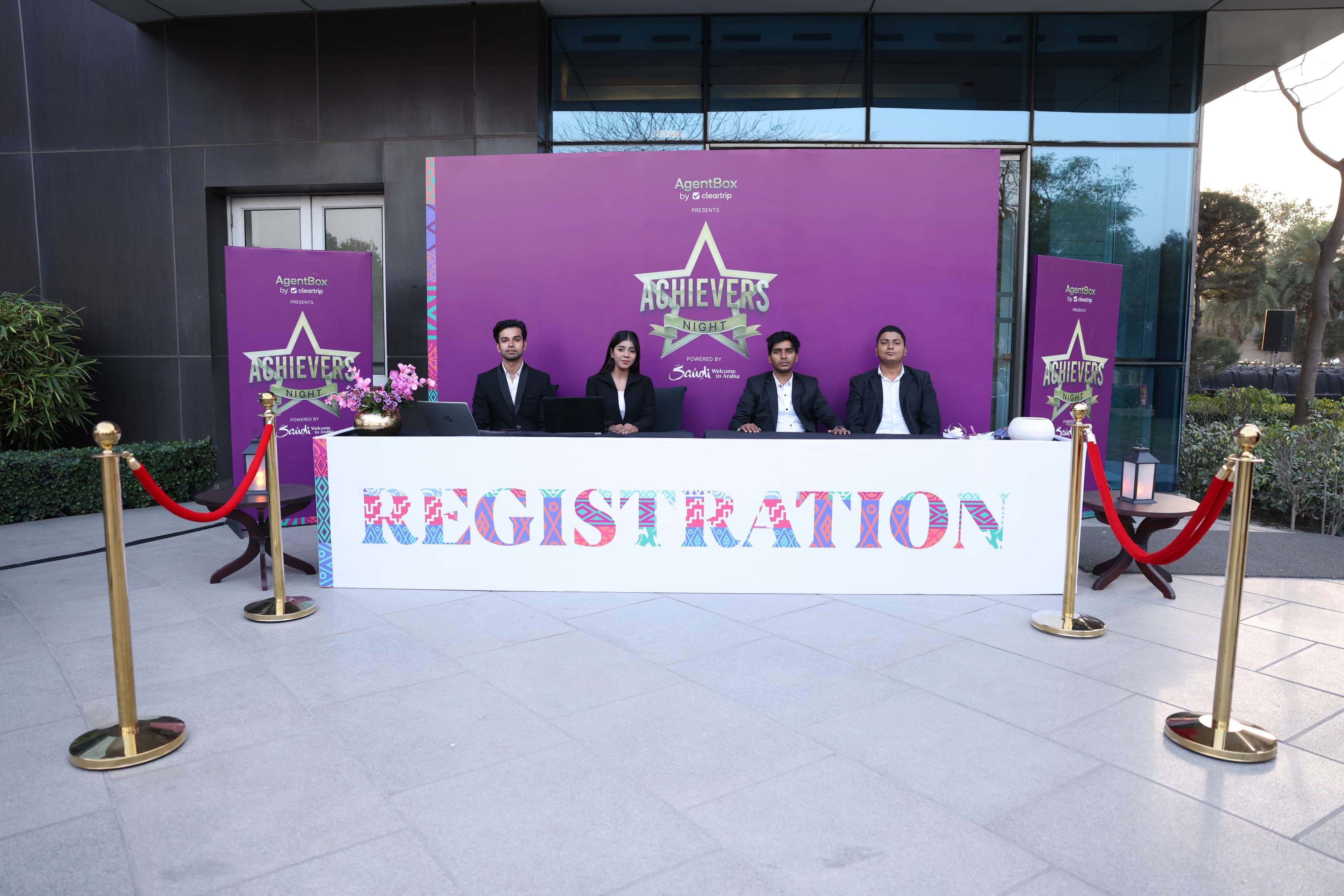 Four hosts seated behind the Achievers Night registration desk, the AgentBox by Cleartrip and 'Powered by Saudi, Welcome to Arabia' branding on the backdrop behind them, red-roped queue stanchions in front