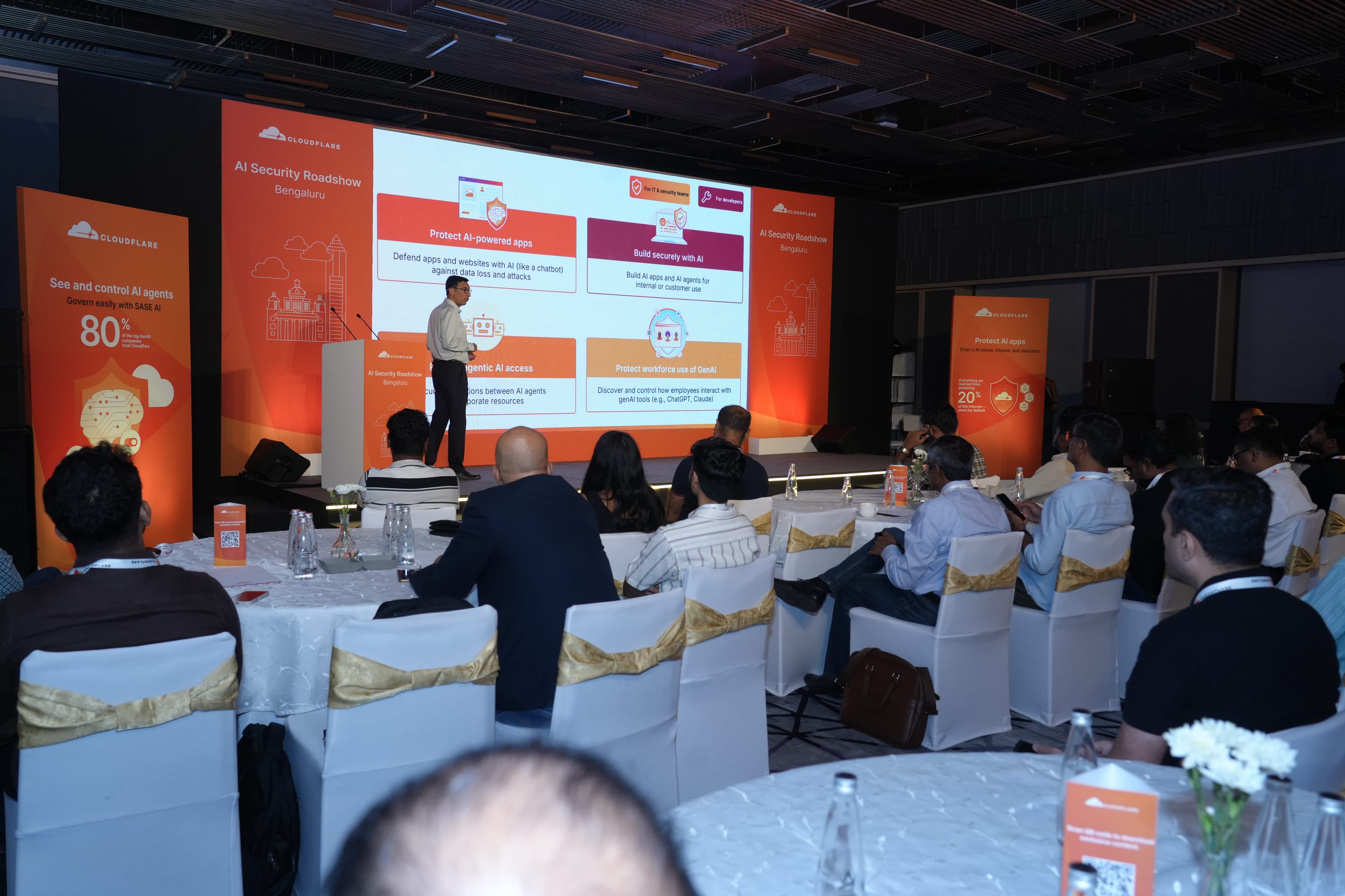 A presenter at the AI Security Roadshow Bengaluru stage walks through four ways to secure AI - protecting AI-powered apps, building securely with AI, governing agentic AI access, and protecting workforce use of GenAI tools like ChatGPT and Claude - each labelled on the screen behind him