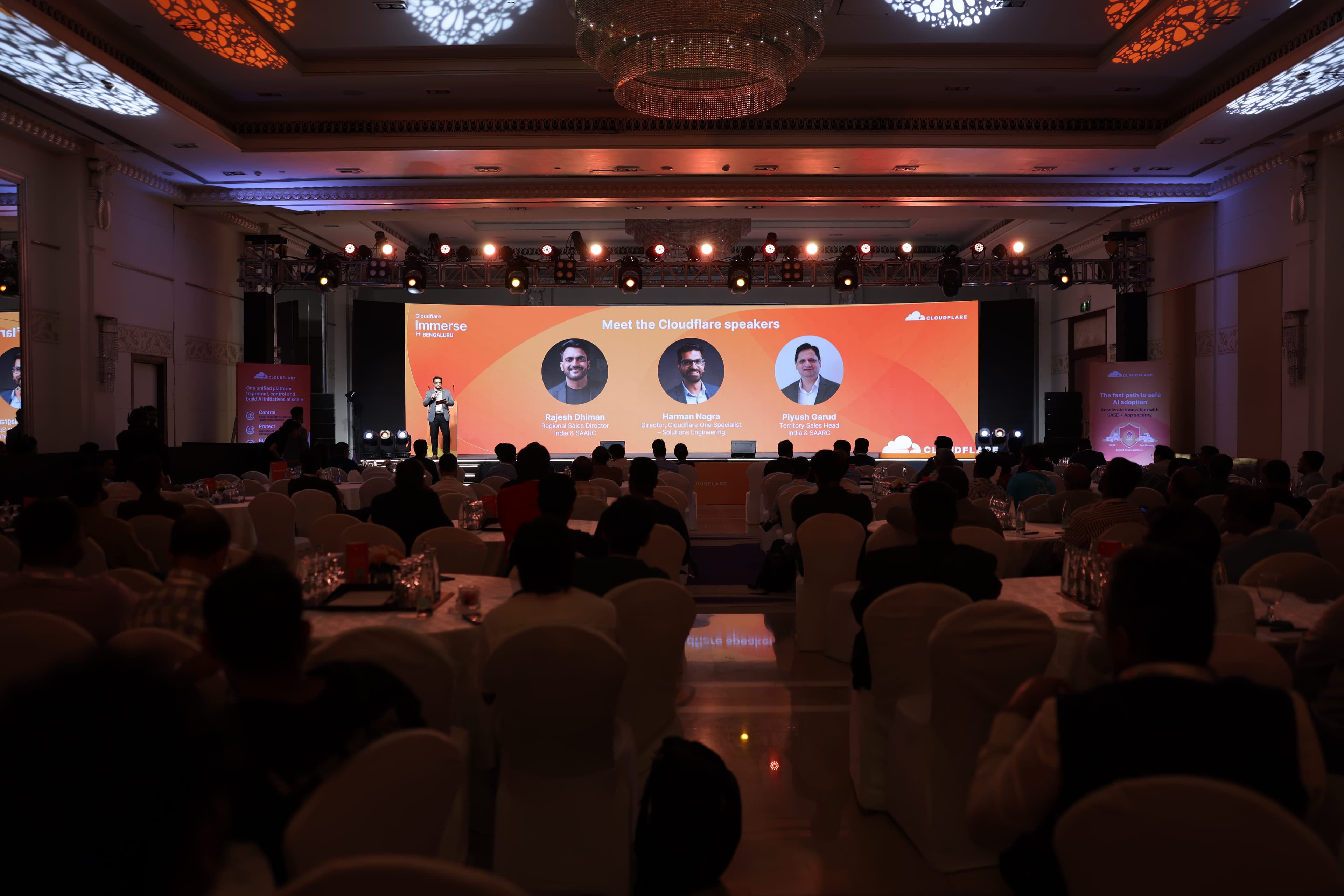 The Cloudflare Immerse hall in Bengaluru at capacity, every round table taken, a speaker on stage introducing the Cloudflare speaker line-up on the wide LED behind him under a full lighting rig