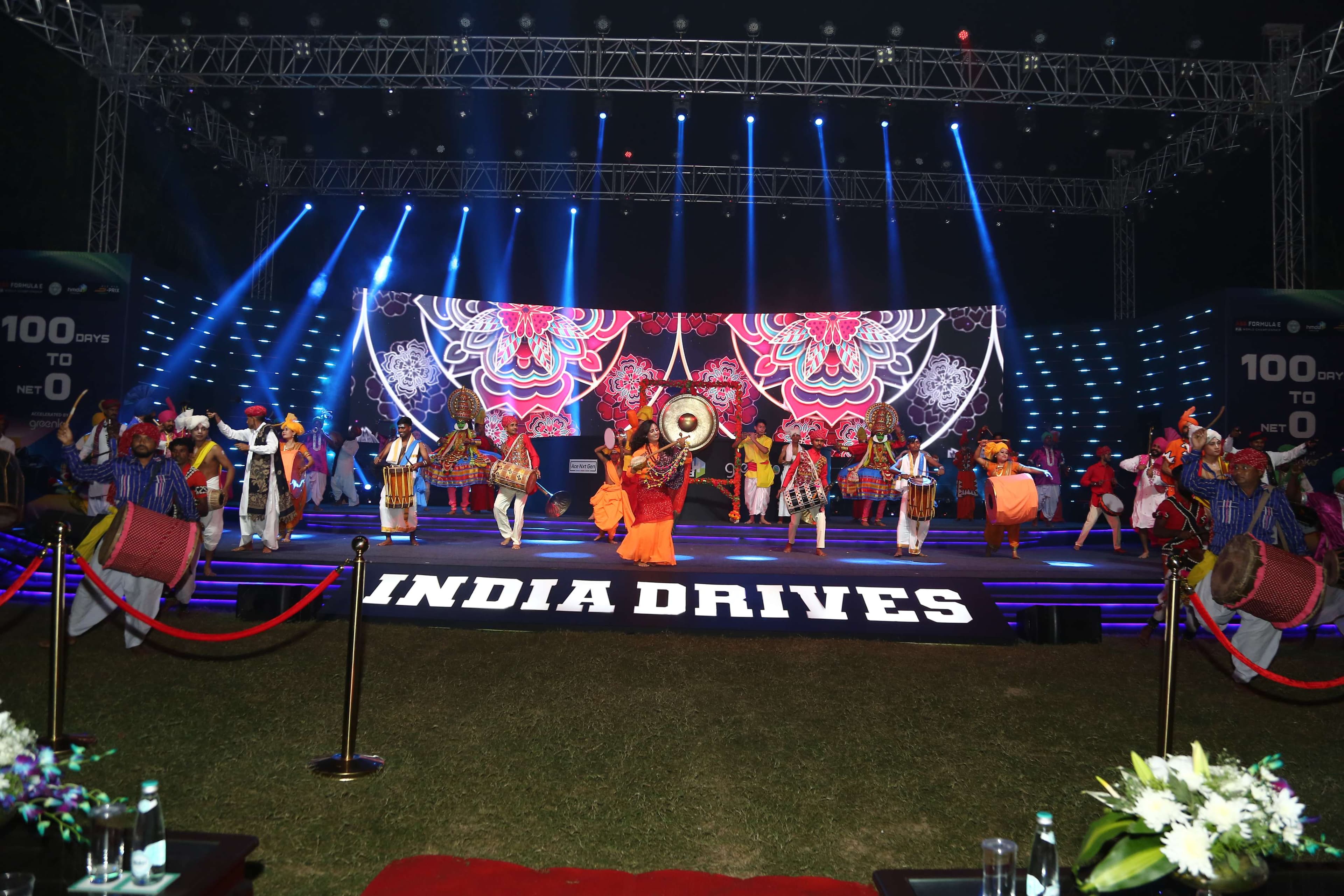The India Drives stage at night, a performer striking a garlanded ceremonial gong flanked by two Kathakali dancers in full costume, 'Formula E', 'hmda' and '100 Days To Net Zero, Accelerated By Greenko' lit on the flanking screens