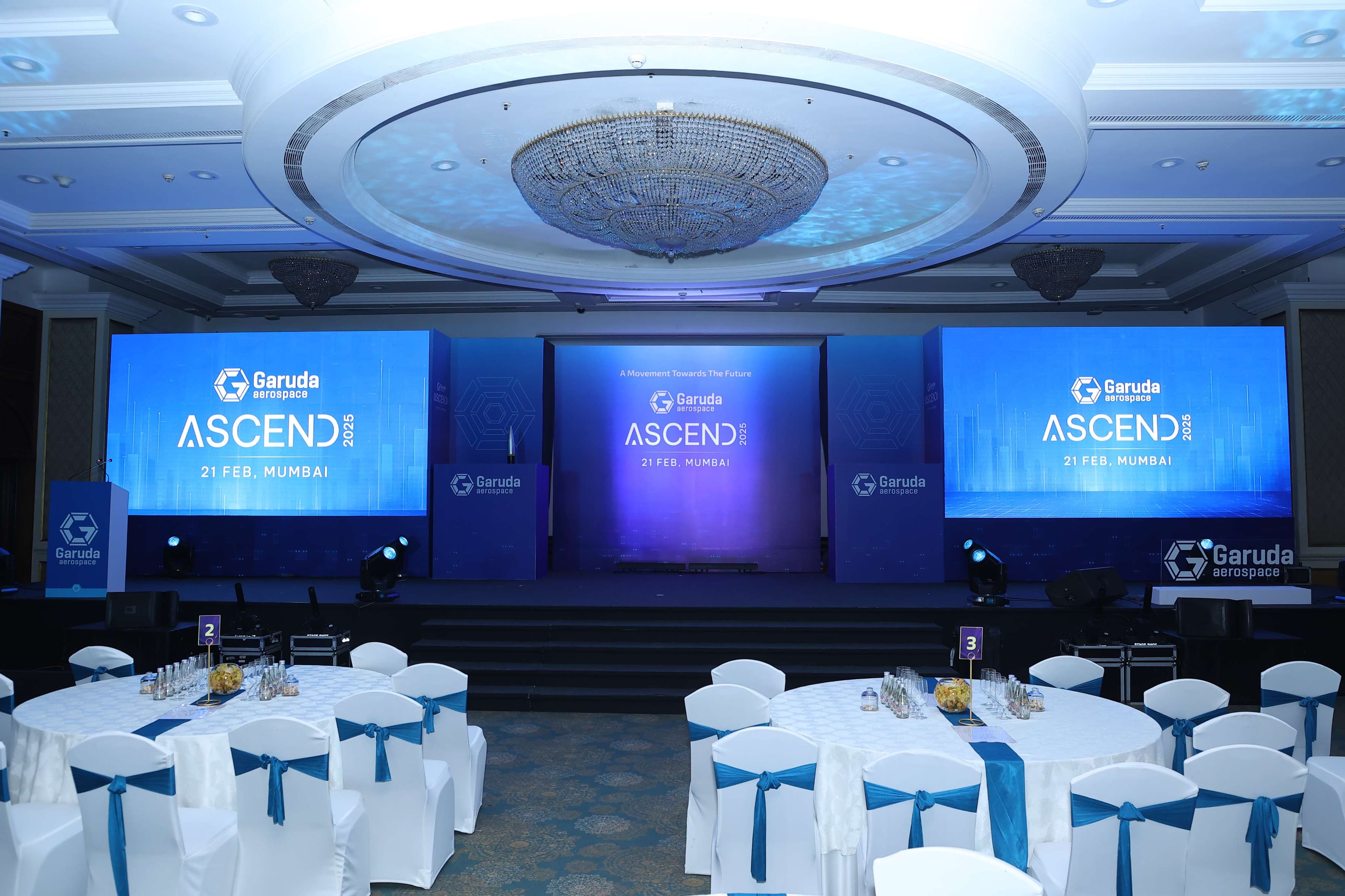 The empty ASCEND 2025 main stage at The Taj Lands End, Mumbai, three screens carrying the Garuda Aerospace ASCEND 2025 branding and the tagline 'A Movement Towards The Future' above rows of blue-draped banquet tables