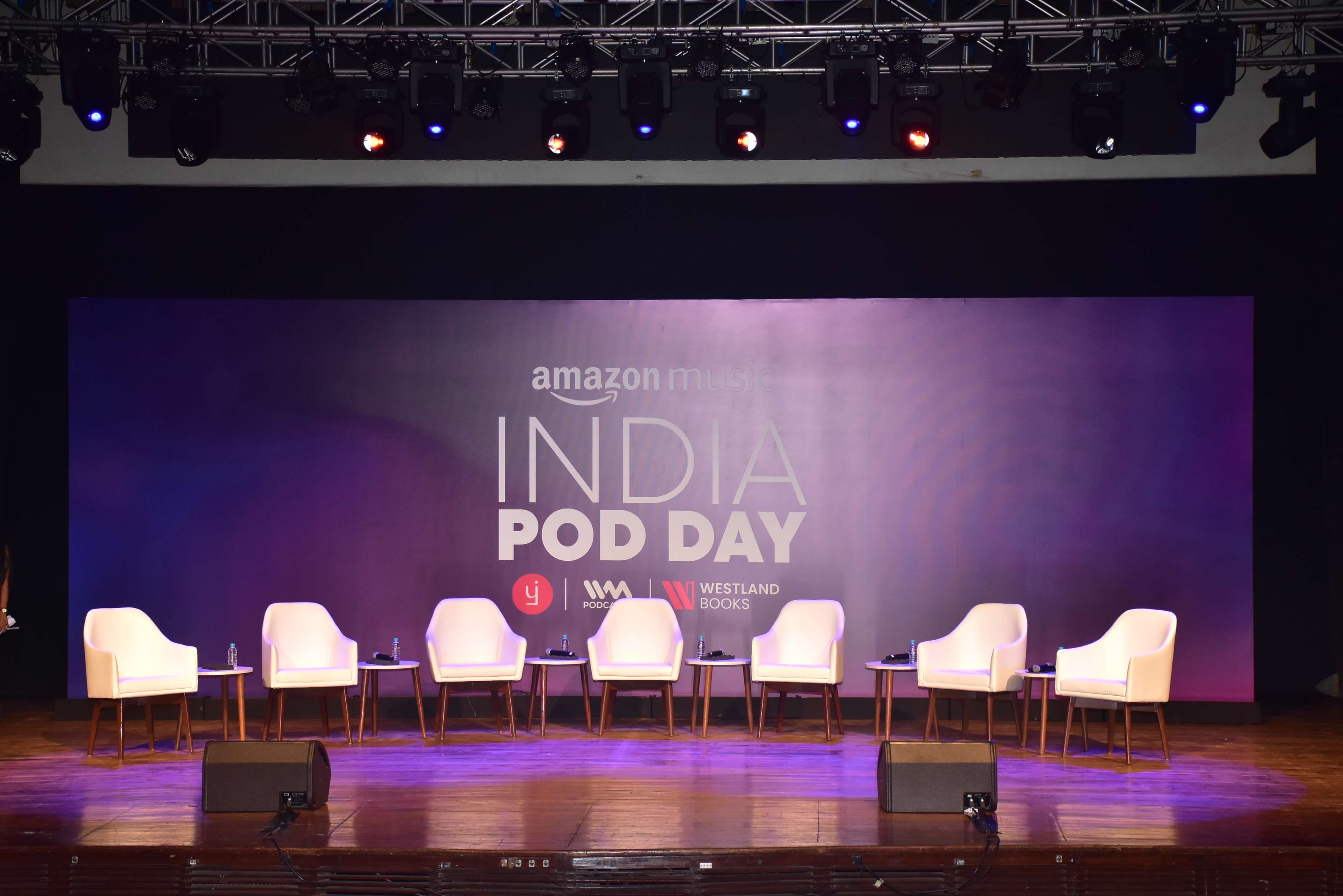 The empty India Pod Day main stage, six chairs and side tables in a row beneath a full lighting truss, the 'amazon music presents India Pod Day' branding lit on the screen behind them