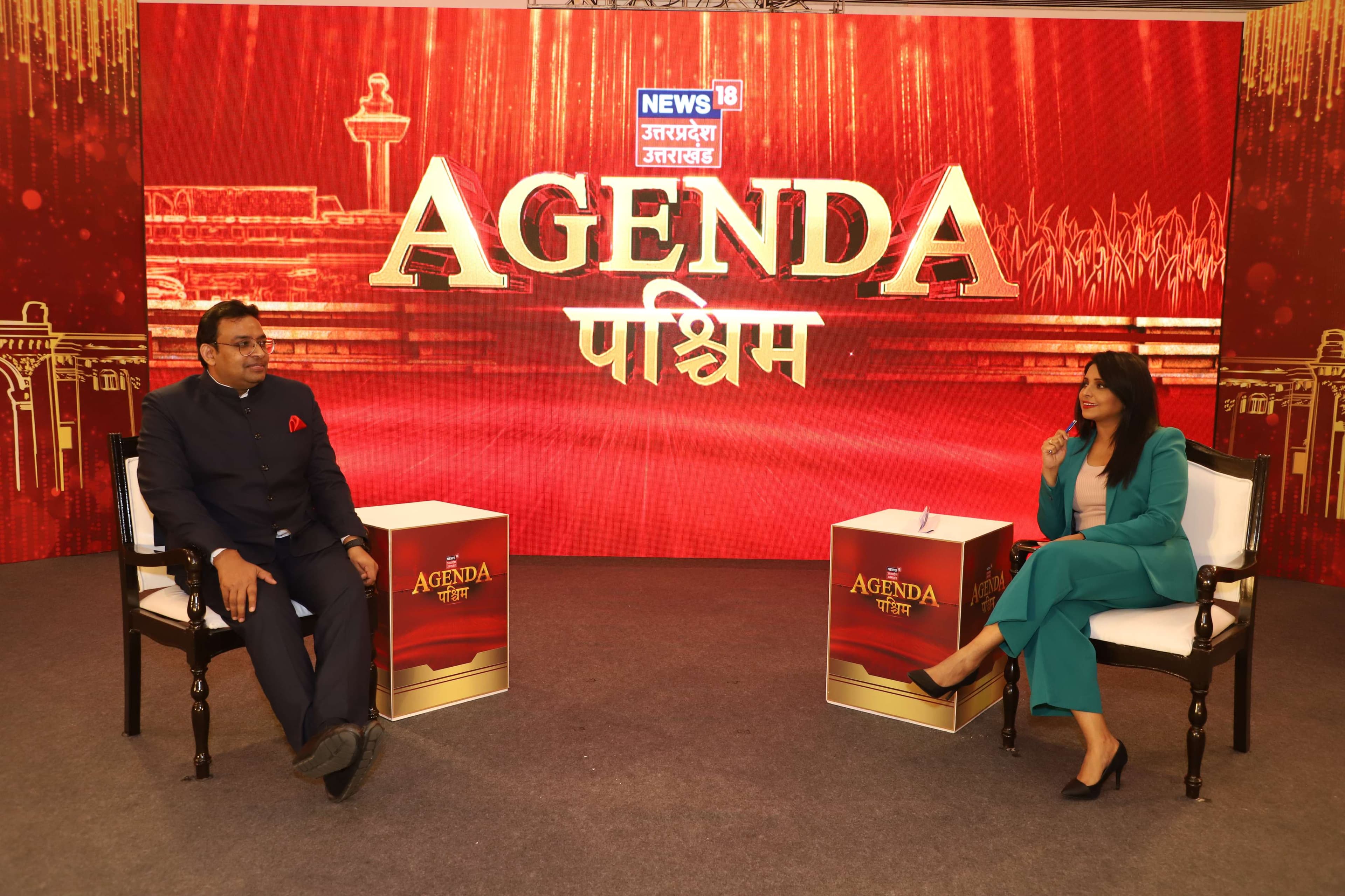 A guest and a News18 anchor in conversation on the Agenda Paschim set, the show's branding and an illustrated monument backdrop lit in red and gold behind them