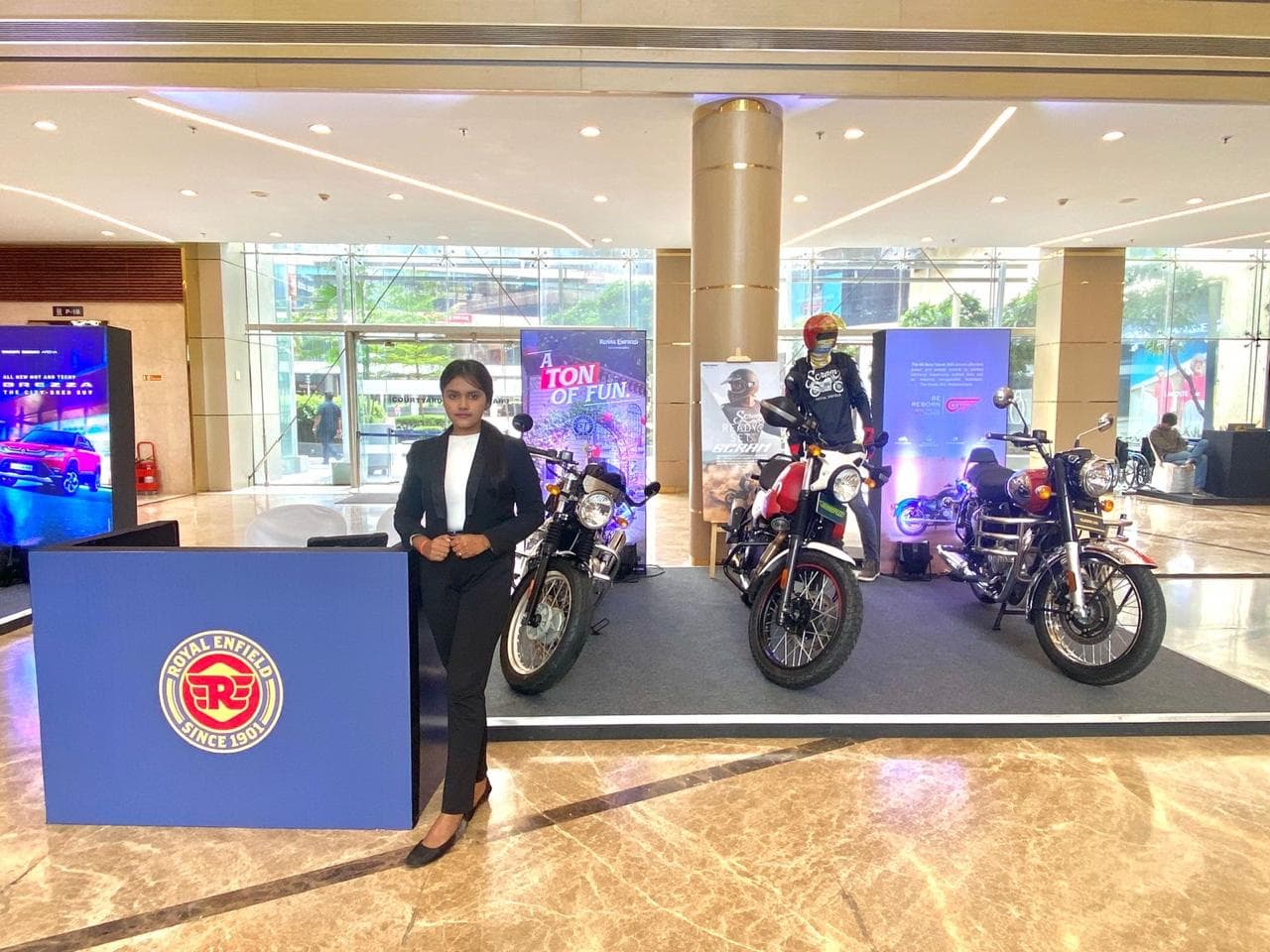 A Royal Enfield display in a shopping mall atrium, three motorcycles lined up on a grey deck behind a navy plinth carrying the brand's logo, a host in a black suit standing at the plinth, 'A Ton Of Fun' and Scram banners and a helmeted mannequin in riding gear behind
